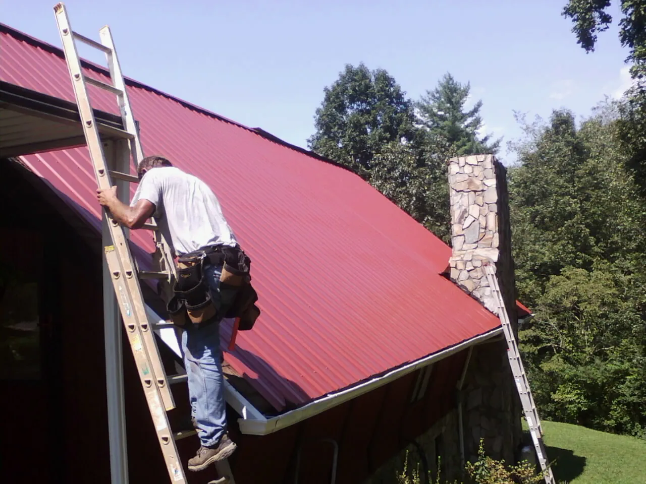 Roofer on a ladder beside a red metal roof and stone chimney — Soffit & Fascia Repair by Roofing Brothers in Cochecton